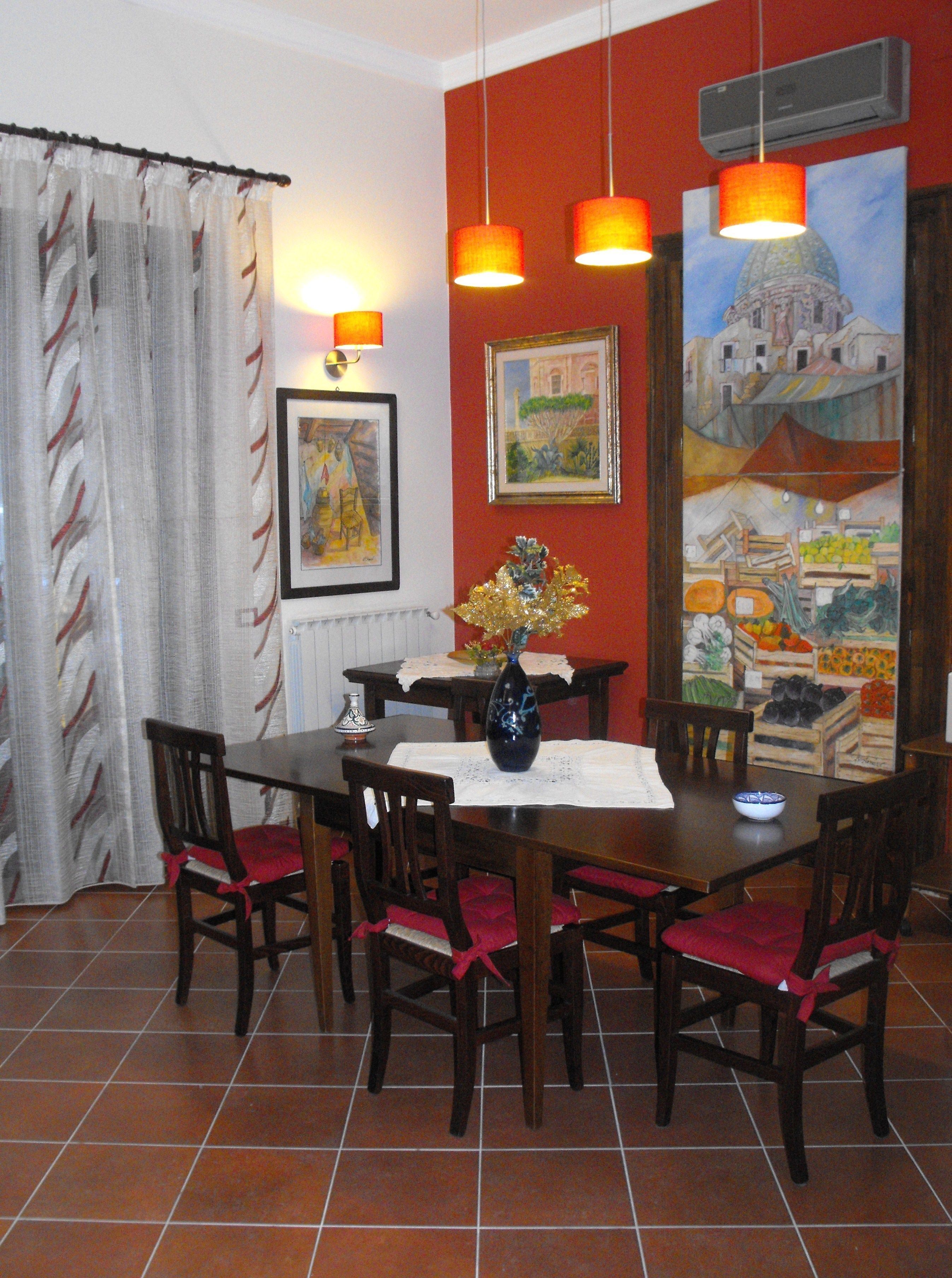 A dining area with a table set for four, surrounded by chairs with red cushions. There are two hanging pendant lights above the table, and a painting depicting a market scene is on the wall.