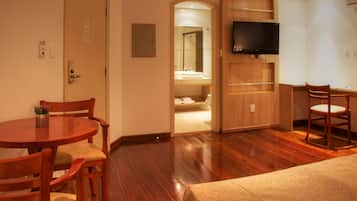 A hotel room with a wooden floor, a round dining table with chairs, a television mounted on the wall, and a bathroom visible through an open door.