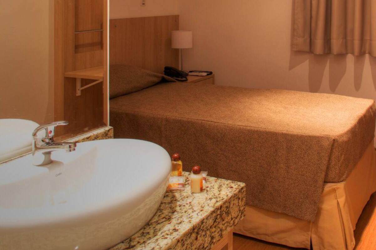 Una habitación de hotel con un lavamanos grande y redondo, una cama con colcha marrón, una mesita de noche con lámpara y un cuadro en la pared.