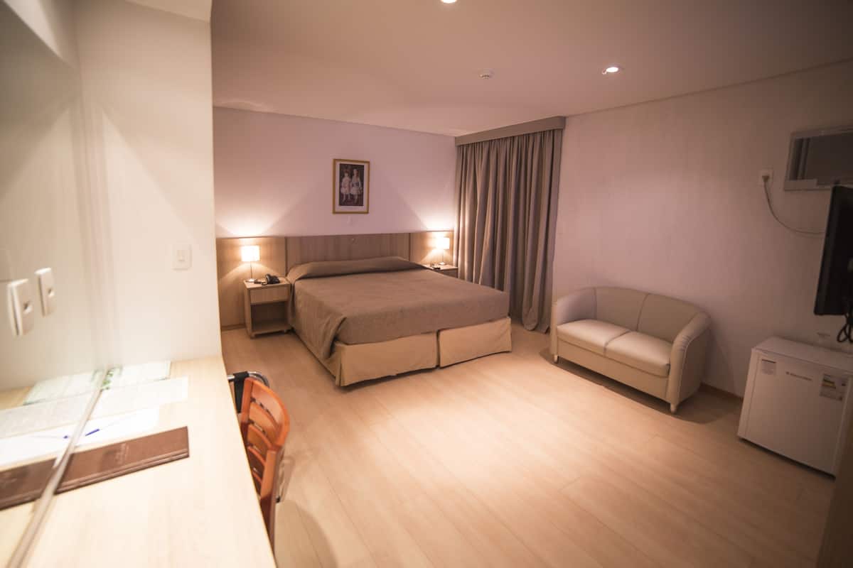 Habitación de hotel con cama, mesitas de noche, una silla, escritorio, televisor y un refrigerador pequeño.