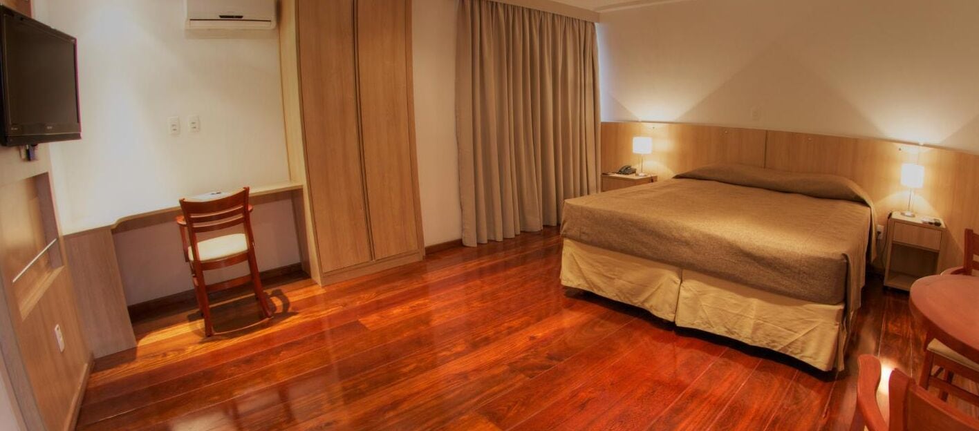 A hotel room with a wooden floor, a bed with a beige bedspread, a desk with a chair, a television, and a window with curtains.