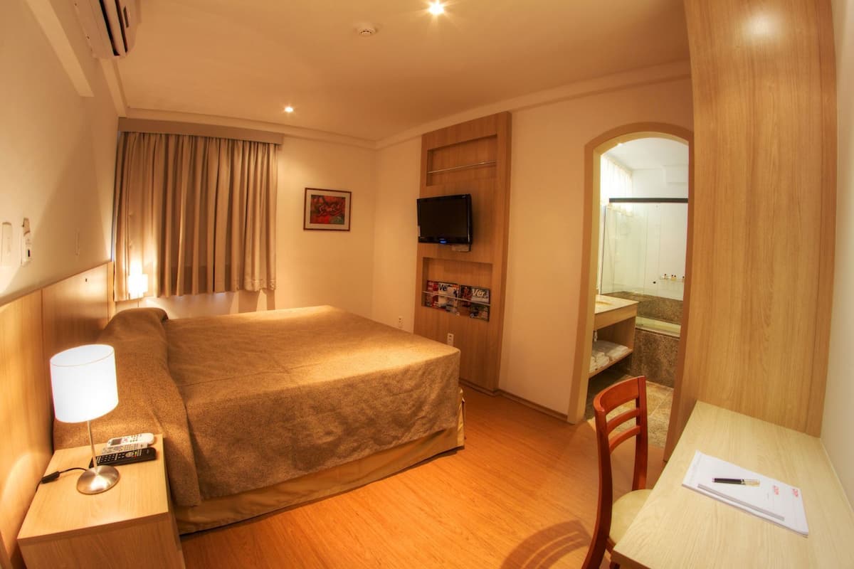 Habitación de hotel con cama, mesita de noche, lámpara, televisor y escritorio con silla.