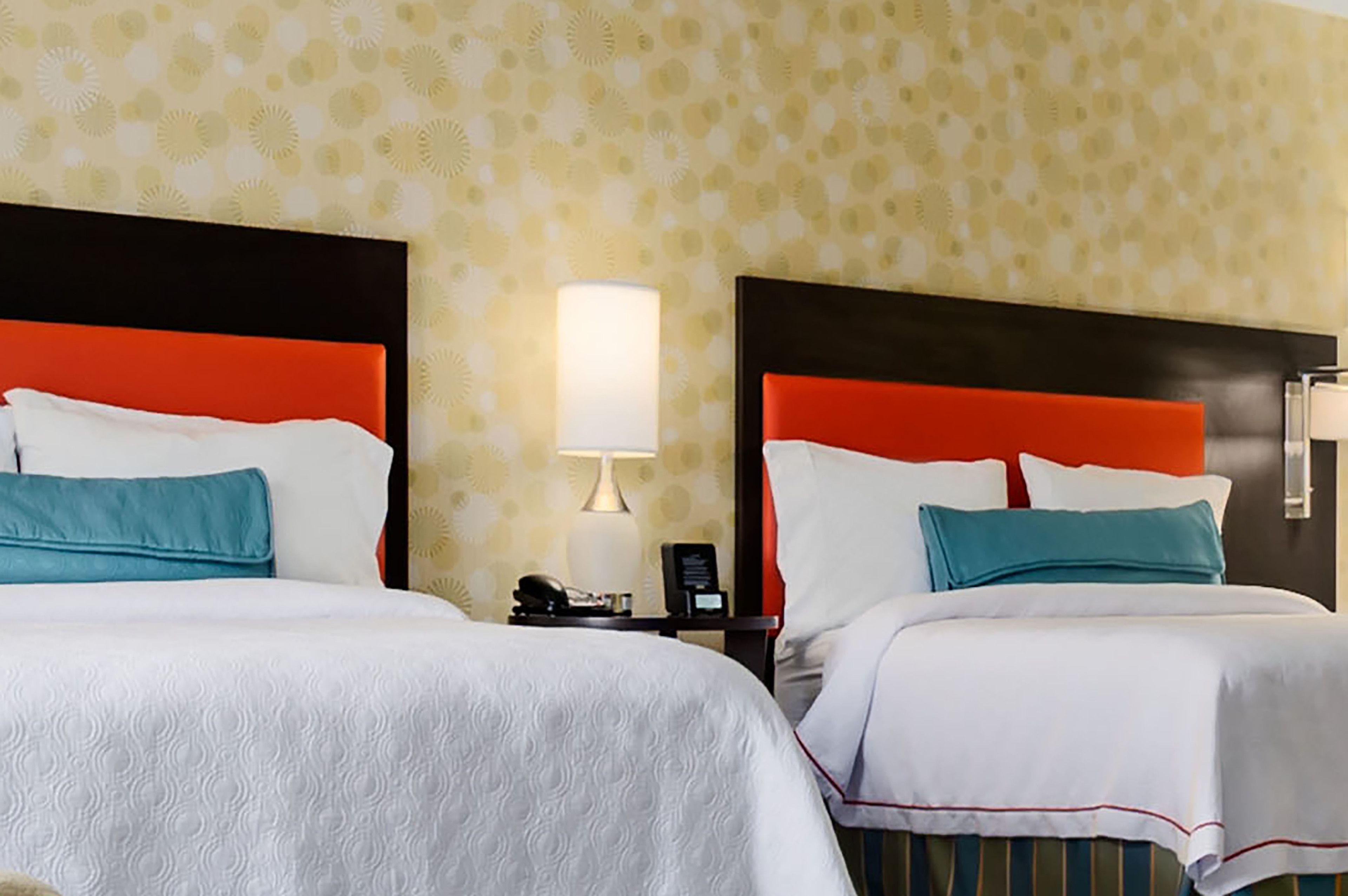 A hotel room with two beds, each with white bedding and teal pillows, and a red headboard. There is a bedside table with a lamp and a phone.