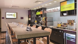 A hotel breakfast area with a long table, chairs, a refrigerator, and a buffet counter.