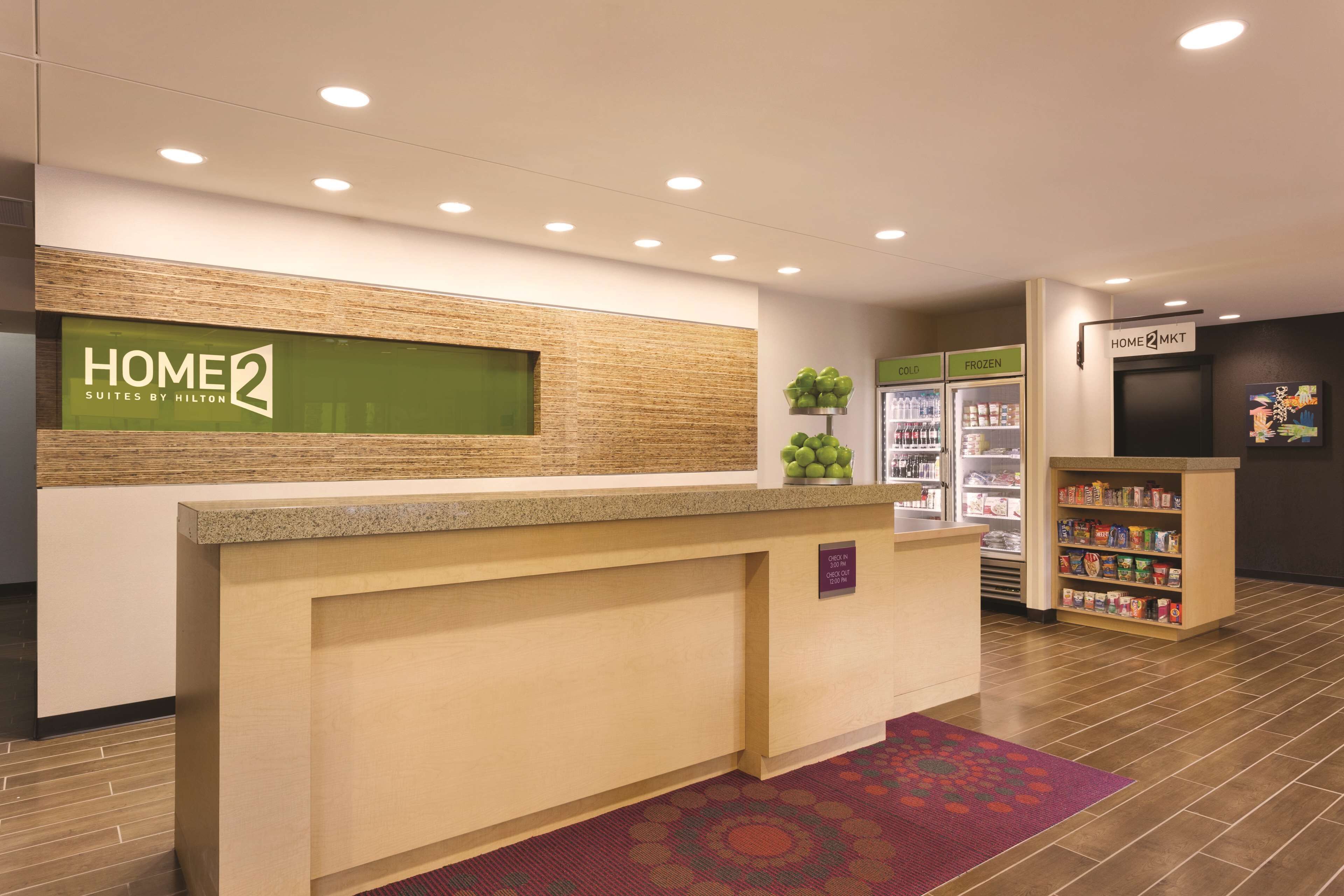 A hotel reception area with a reception desk, a green sign reading "HOME 2 Suites by Hilton," a refrigerator with a "FROZEN" sign, and a self-service kiosk.