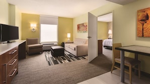 A hotel room with a television, sofa, ottoman, dining table, and a painting on the wall.