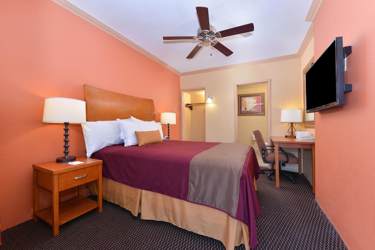 A hotel room with a bed, bedside tables, a desk, a chair, a ceiling fan, and a TV.