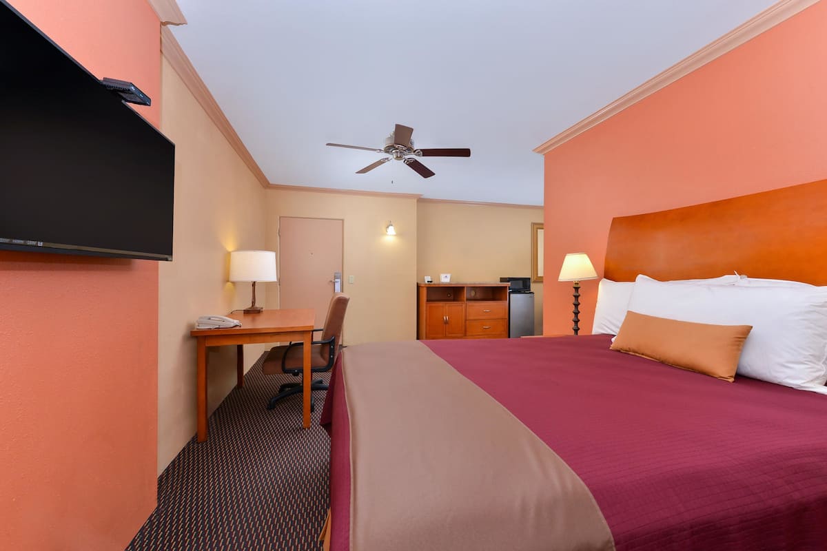 A hotel room with a bed, desk, chair, television, ceiling fan, and a small kitchenette.