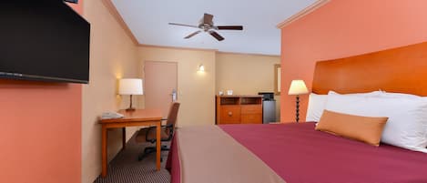 A hotel room with a bed, desk, chair, television, ceiling fan, and a small kitchenette.