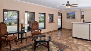 A hotel lobby with a reception desk, a glass-top table, two upholstered chairs, a lamp, and a patterned rug.