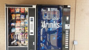 A vending machine with snacks and a drink vending machine labeled "COLD drinks" next to a brick wall.