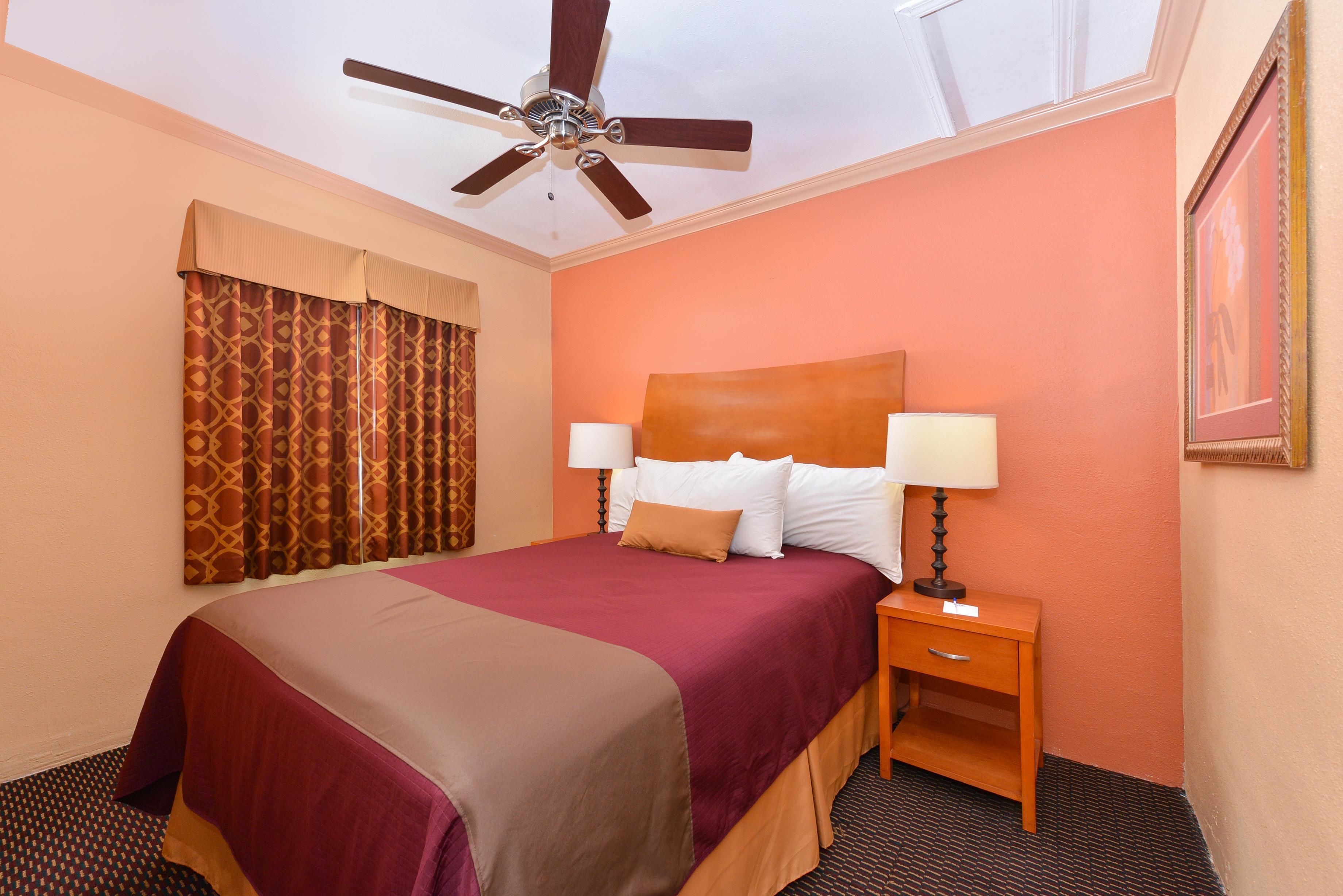 A hotel room with a bed, two bedside lamps, a nightstand, a ceiling fan, and a window with curtains.