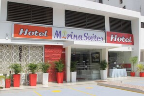Cet hôtel présente une façade moderne, avec une enseigne bien visible indiquant « MarinaSuites » et une autre, plus simple, indiquant « Hôtel ». Plusieurs plantes en pot sont disposées devant l’entrée.