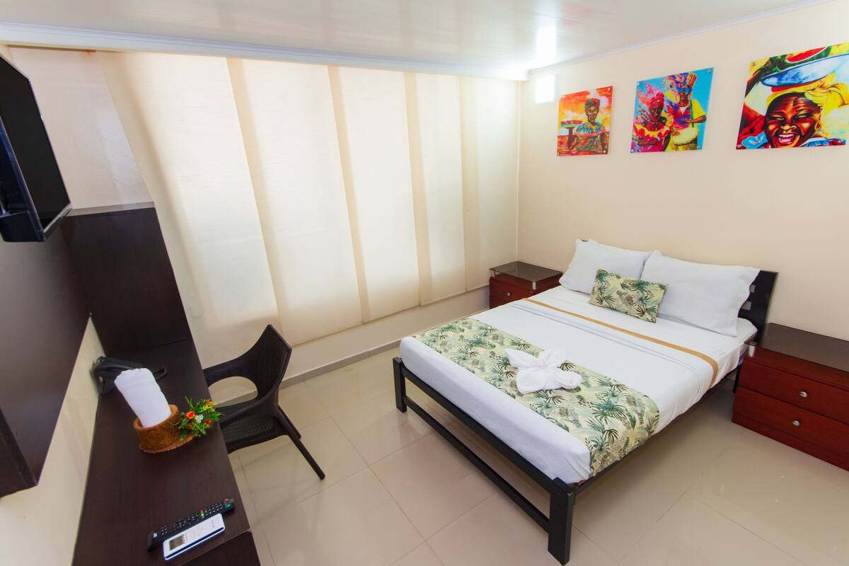 A hotel room with a bed, a desk with a chair, a TV, and artwork on the wall.