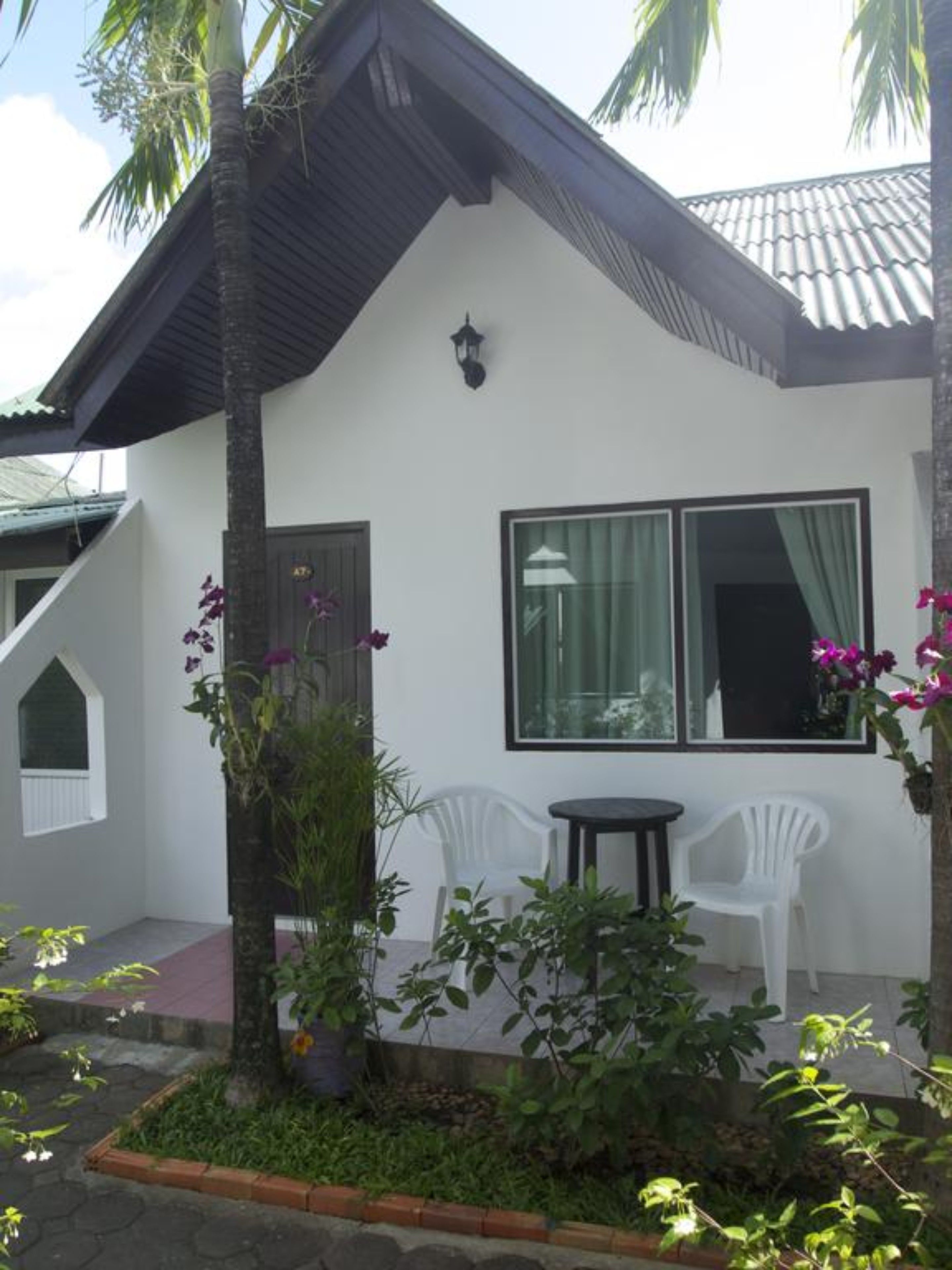 A white building with a dark roof, a large window, and two white chairs outside.
