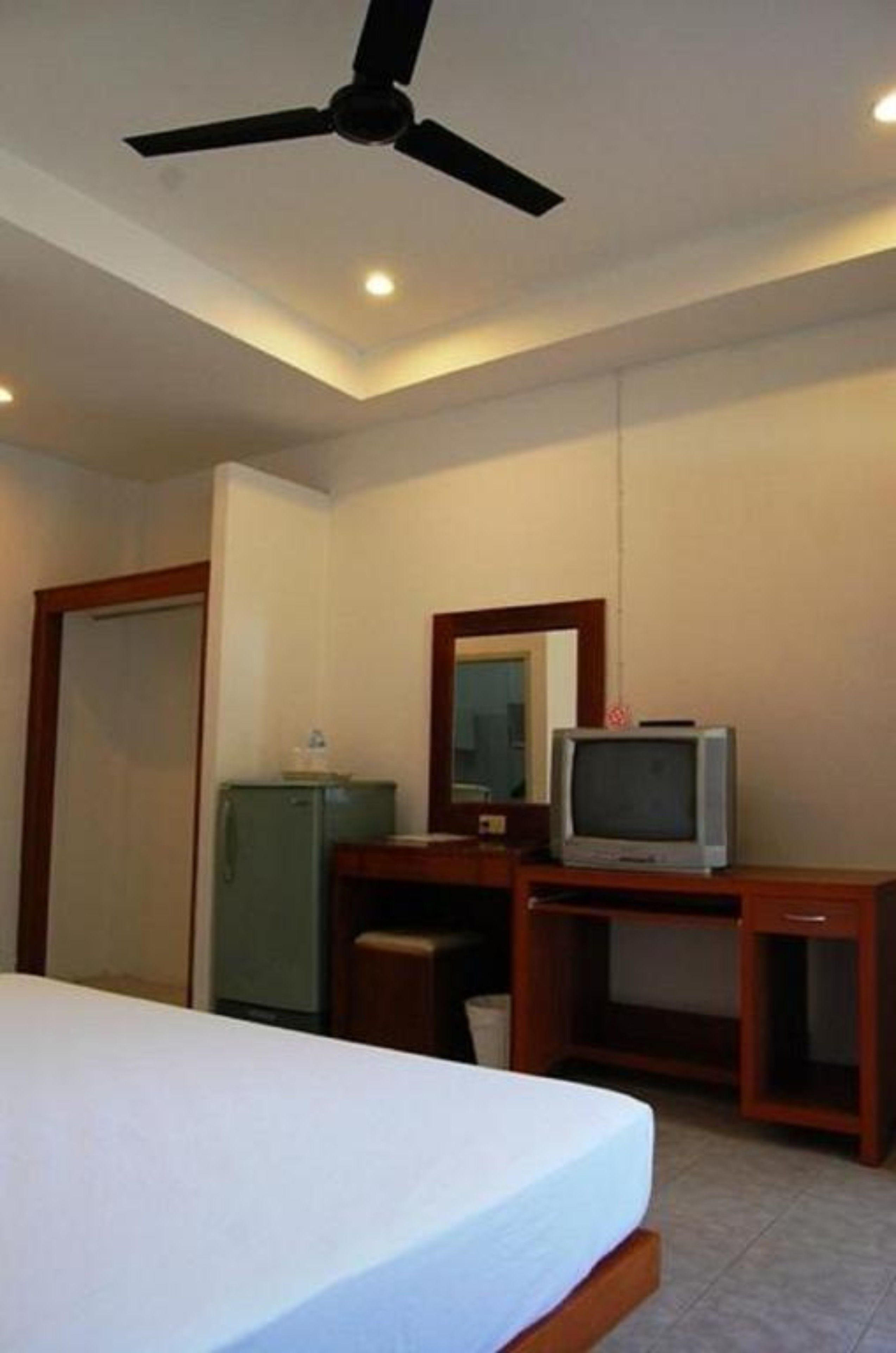 A hotel room with a bed, a desk with a television, a mirror, a ceiling fan, and a small refrigerator.