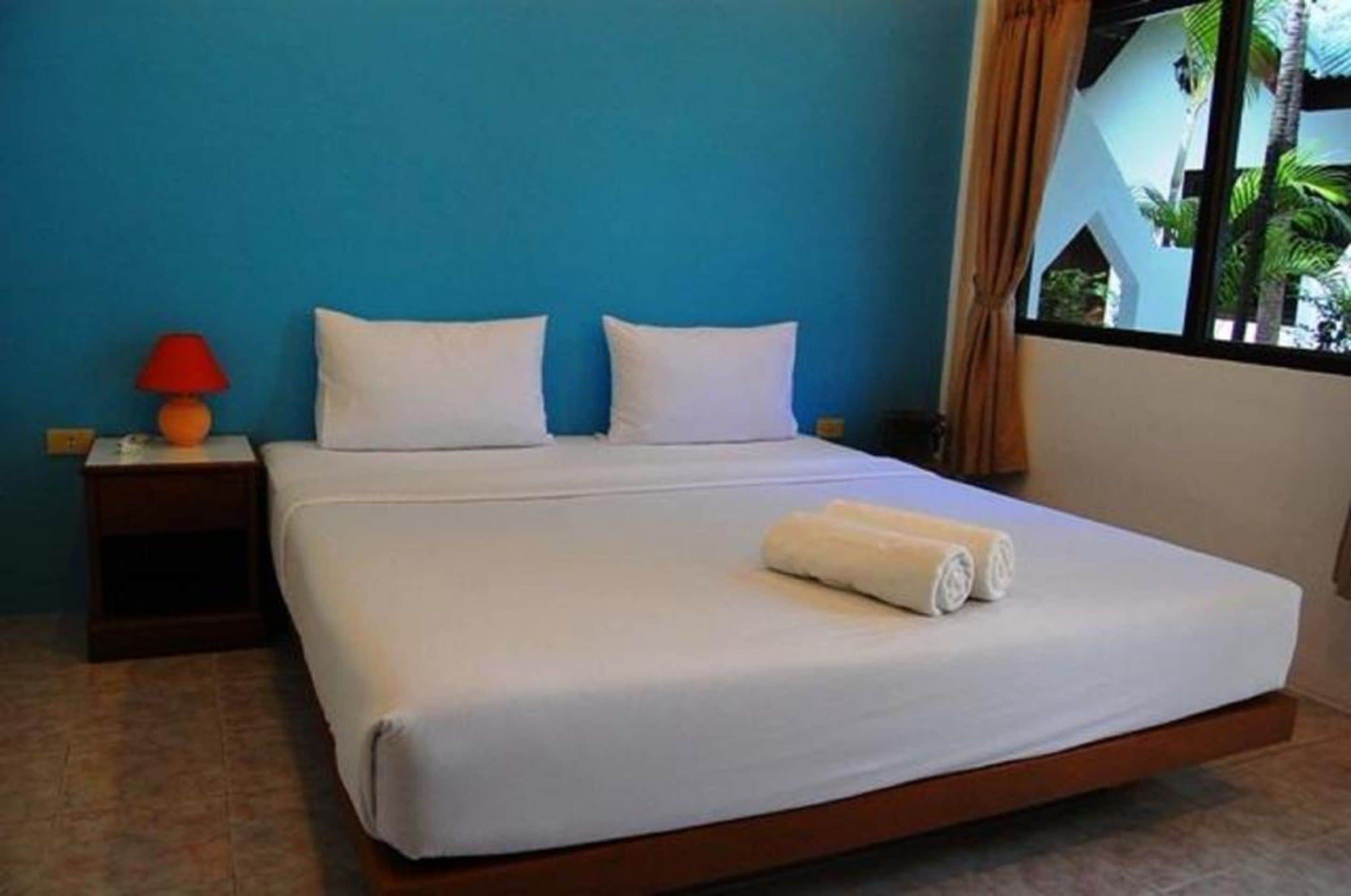 A single bed with white linens, a red lamp on a nightstand, and a window with curtains.