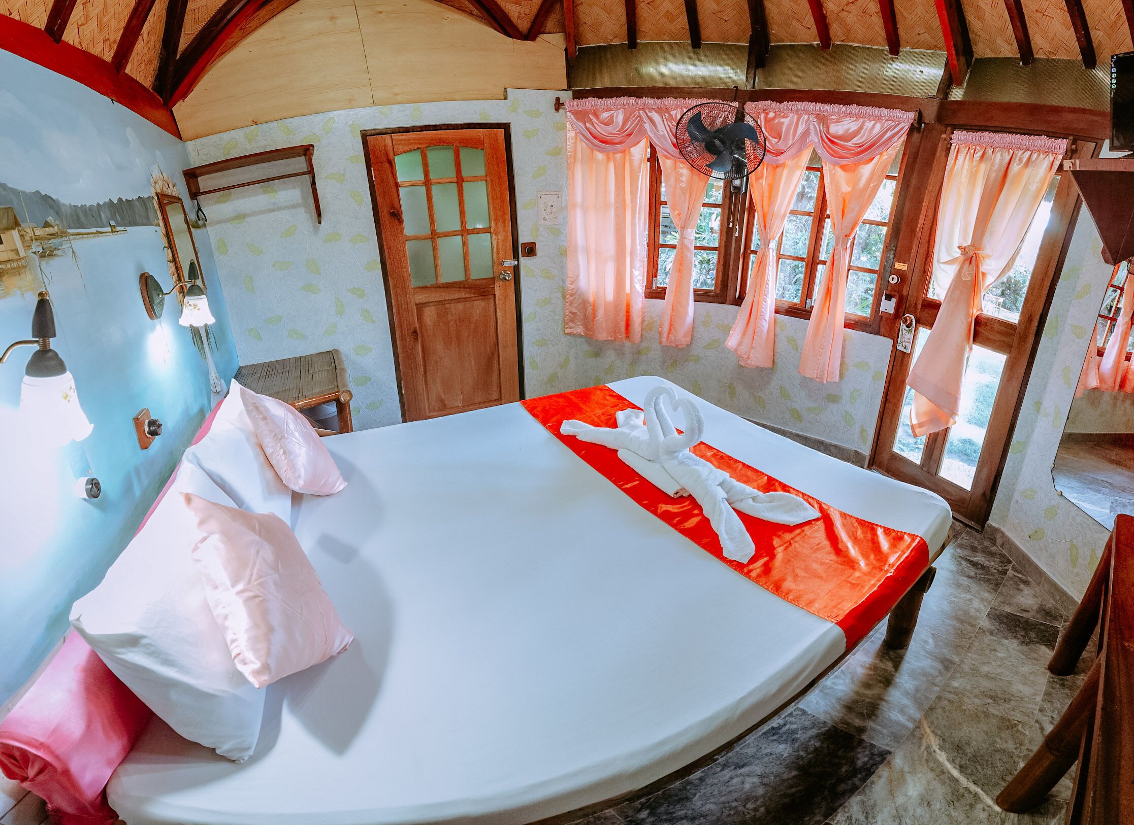 Una habitación con una cama, una puerta de madera, un ventilador y cortinas.