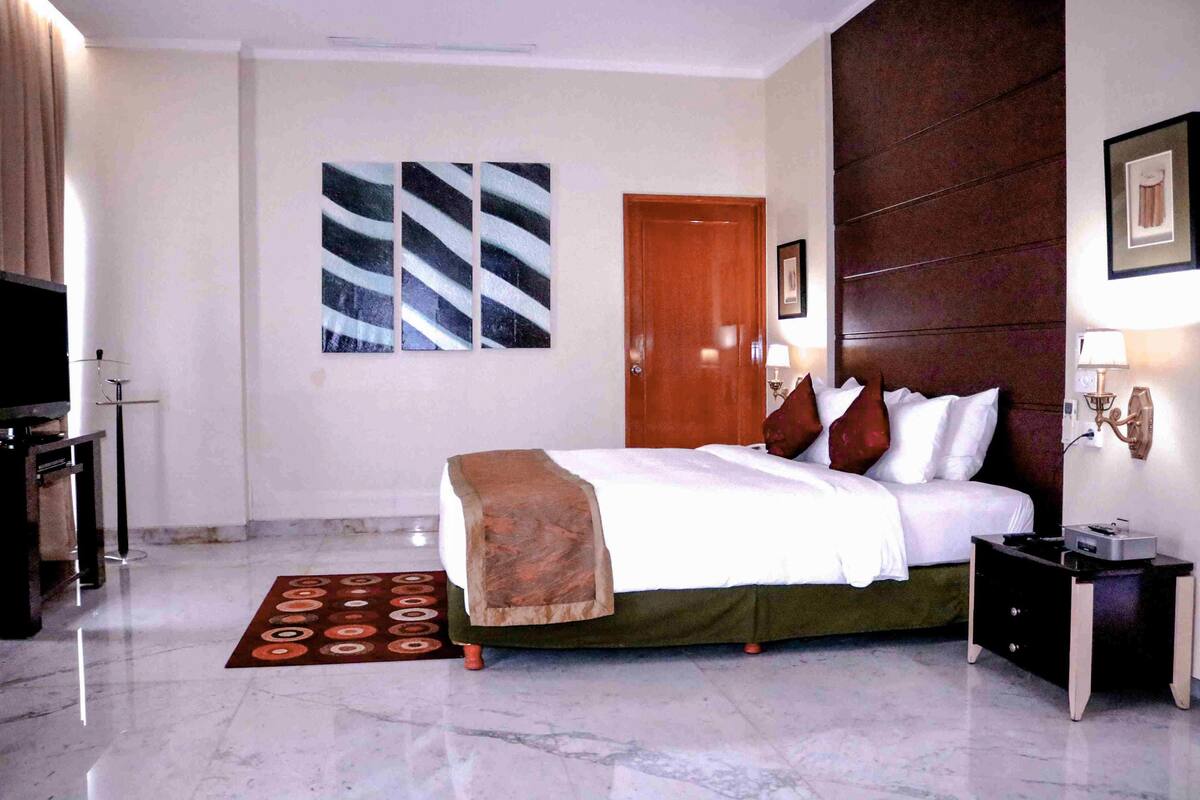 A hotel room with a bed, a wooden door, a nightstand, a lamp, and a wall-mounted picture.
