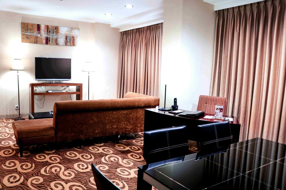 A hotel room with a flat-screen TV, a sofa, a dining table with chairs, and a patterned carpet.