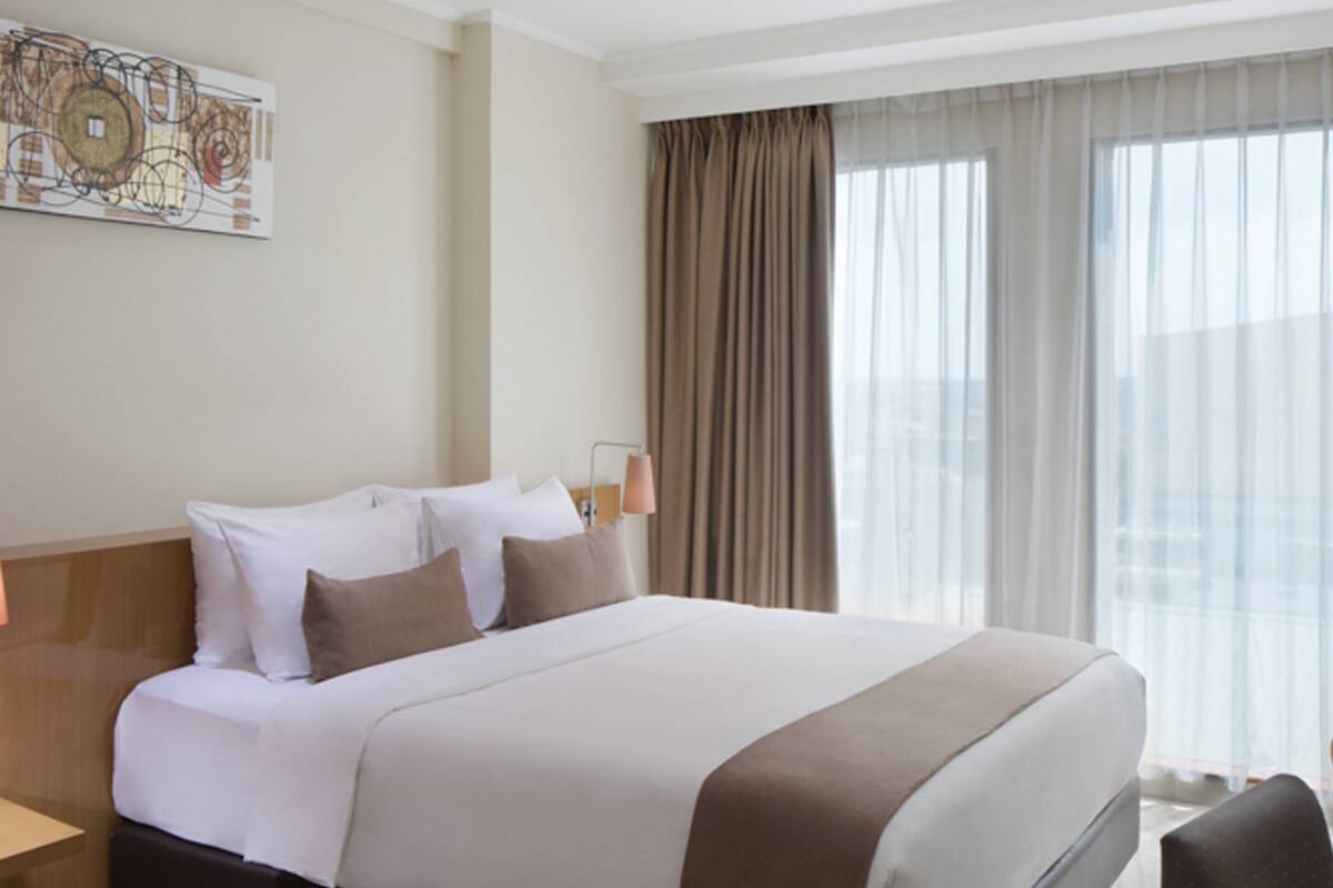 A hotel room with a large bed, a nightstand, a lamp, a chair, and a window with curtains.
