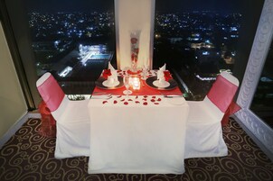 Una mesa para dos adornada con pétalos de rosa, una vela y un florero con una flor roja.