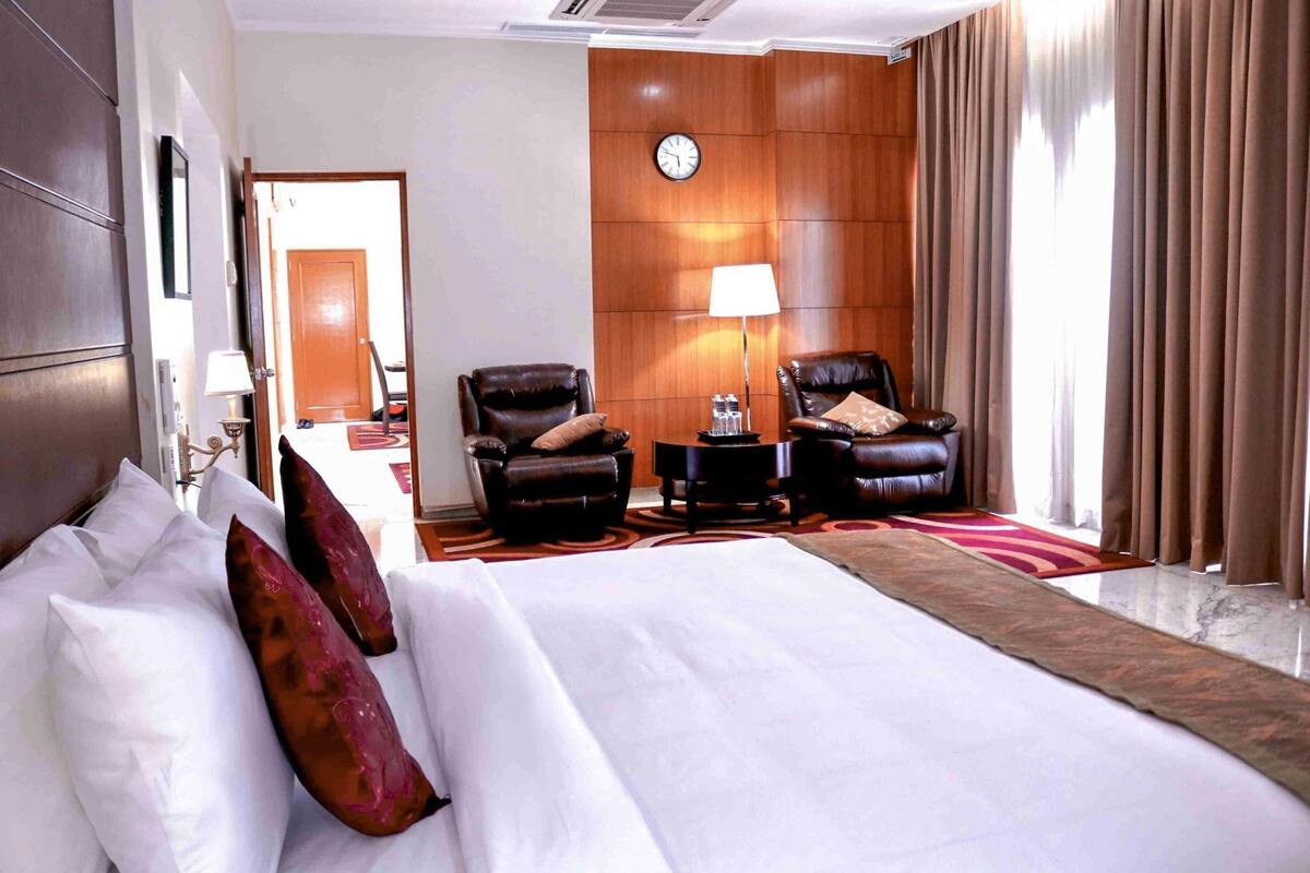 A hotel room with a large bed, two leather armchairs, a side table with a lamp, a clock on the wall, and a view of the outside through a window with curtains.