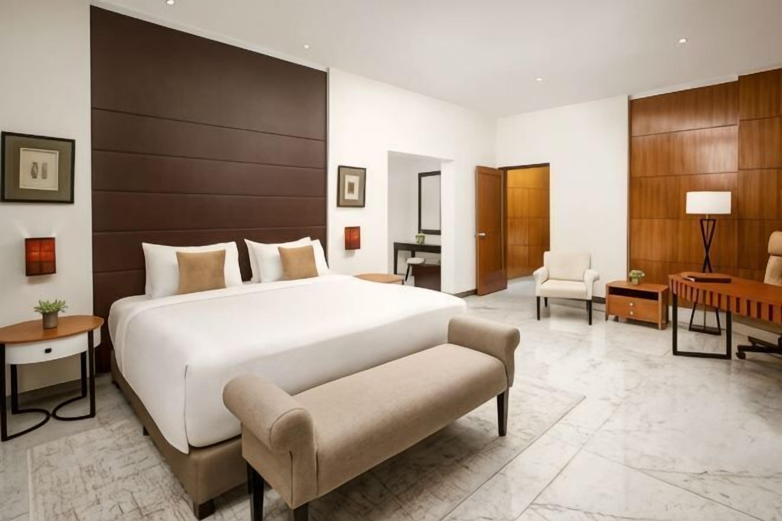 A hotel room with a large bed, a desk, a chair, a bench, a lamp, and a mirror.
