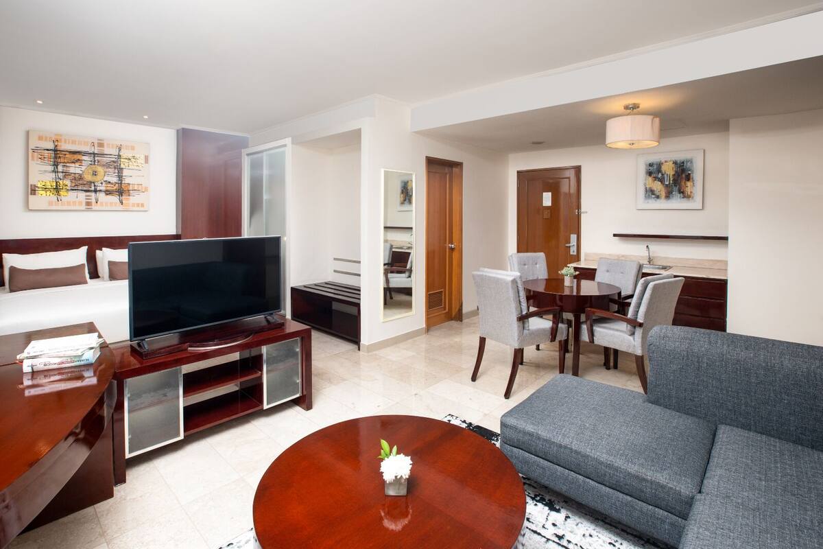 A modern hotel room with a flat-screen TV, a sofa, a dining table with chairs, and a small coffee table.