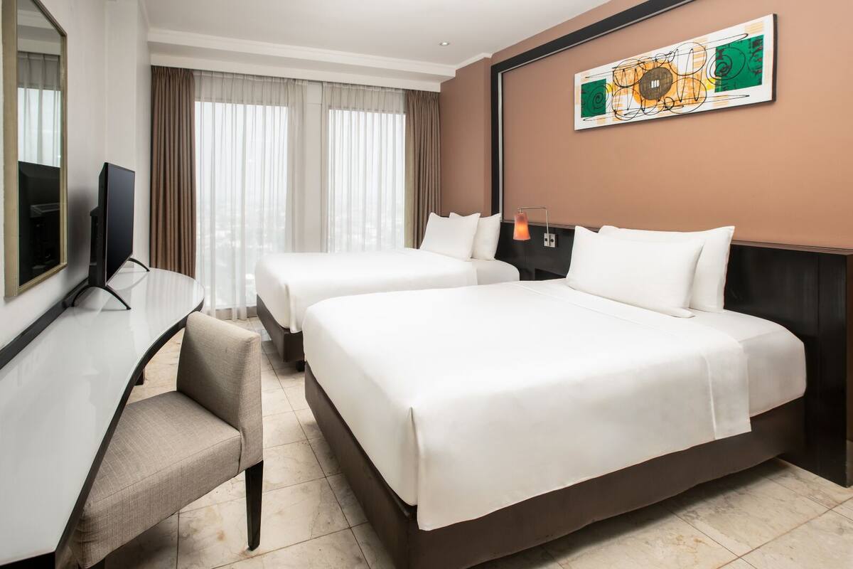 A hotel room with two beds, a desk, a television, and a painting on the wall.