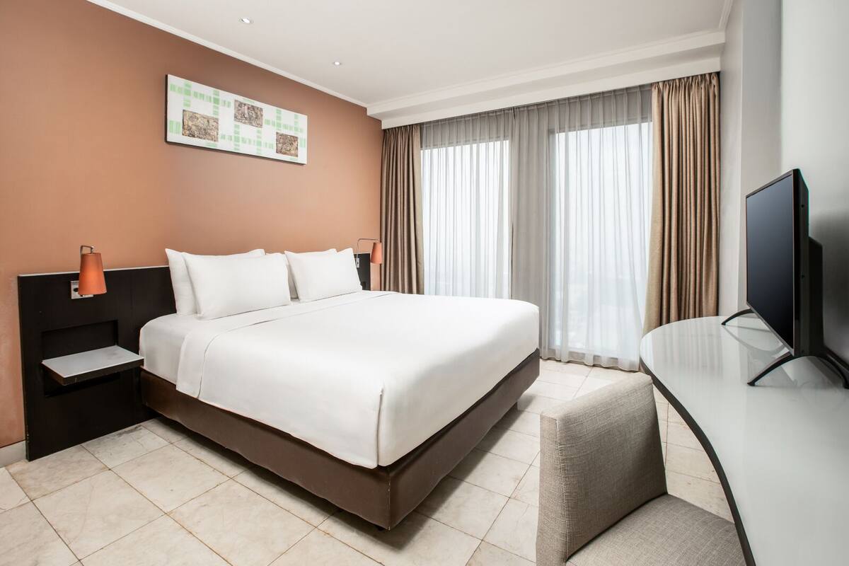 A modern hotel room with a large bed, a desk with a chair, a flat-screen TV, and a window with curtains.