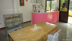 A reception area with a pink counter, a wooden table with a decorative item, a glass display case with snacks, and a window with a view of greenery. - Amerson Pension Place - Hostel (Puerto Princesa)