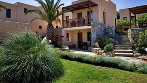 A modern two-story building with a balcony, stone accents, and a well-maintained garden with palm trees and shrubs. - Ida Village I & II (Hersonissos)