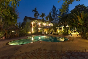 A swimming pool with a tiled deck, surrounded by lush greenery and a building with illuminated windows. - Ladda Resort (Takua Pa)