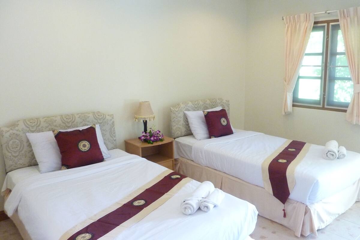 Quarto de hotel com duas camas de solteiro, cada uma com travesseiro, mesa de cabeceira, abajur e janela com cortinas.