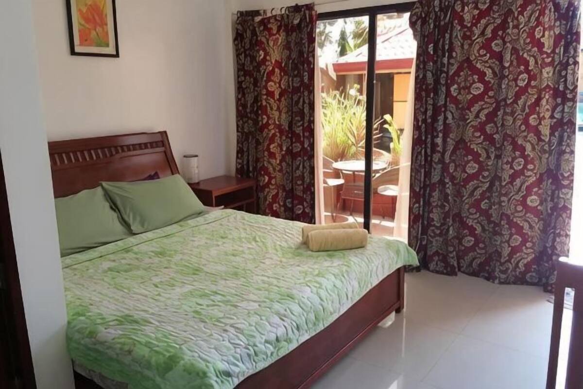 A bedroom with a bed, a nightstand, a chair, and a view of an outdoor area with plants.