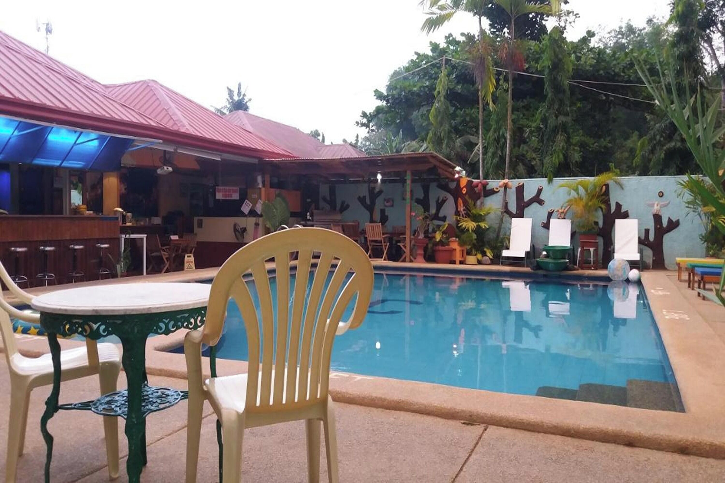 A pool area with a red-roofed building, outdoor seating, and tropical plants.