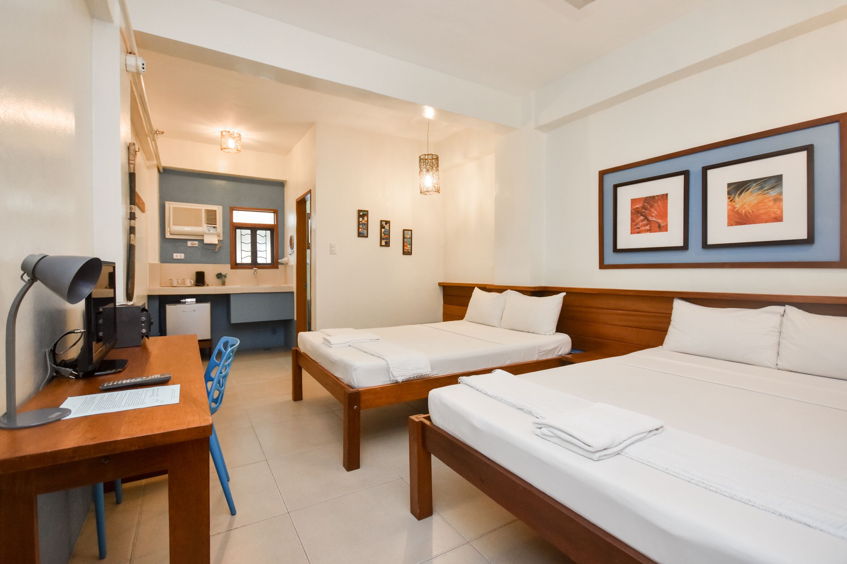 Habitación de hotel con dos camas, un escritorio con lámpara, una silla, una kitchenette pequeña y obras de arte en las paredes.