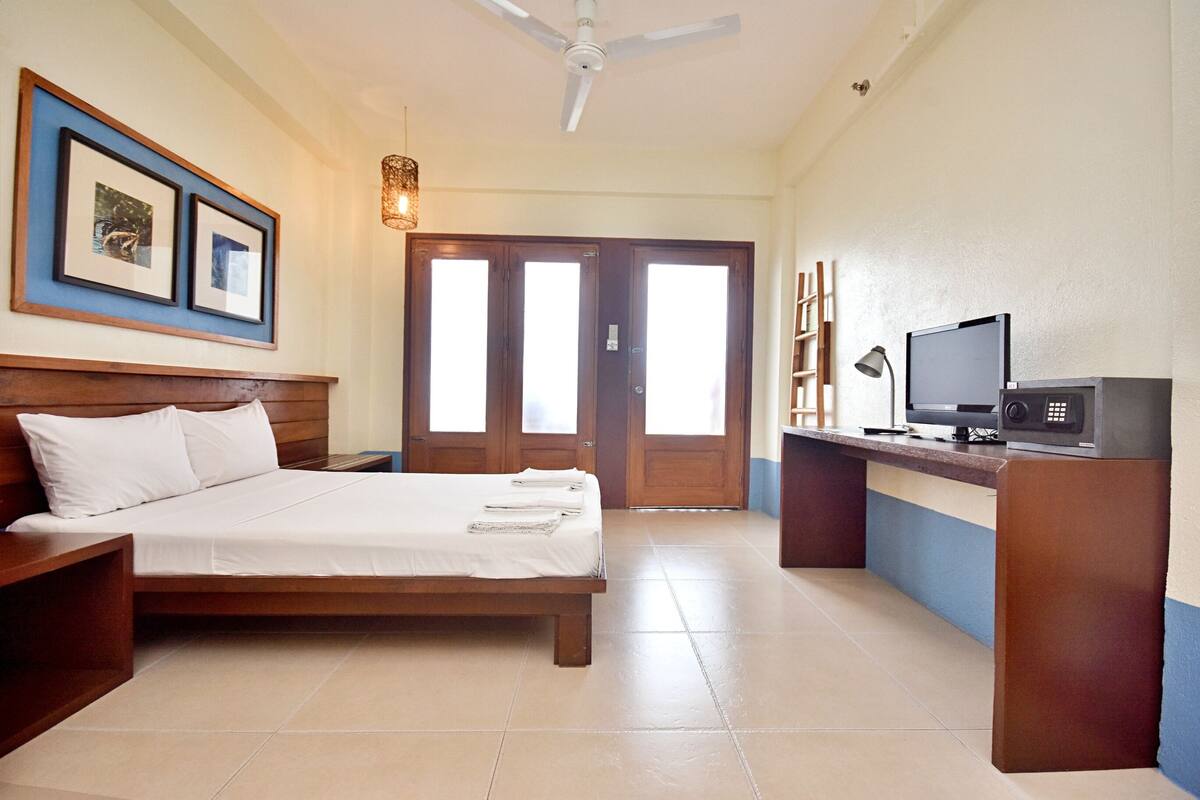 Una habitación de hotel con una cama, un escritorio con computadora, un ventilador de techo y cuadros en la pared.