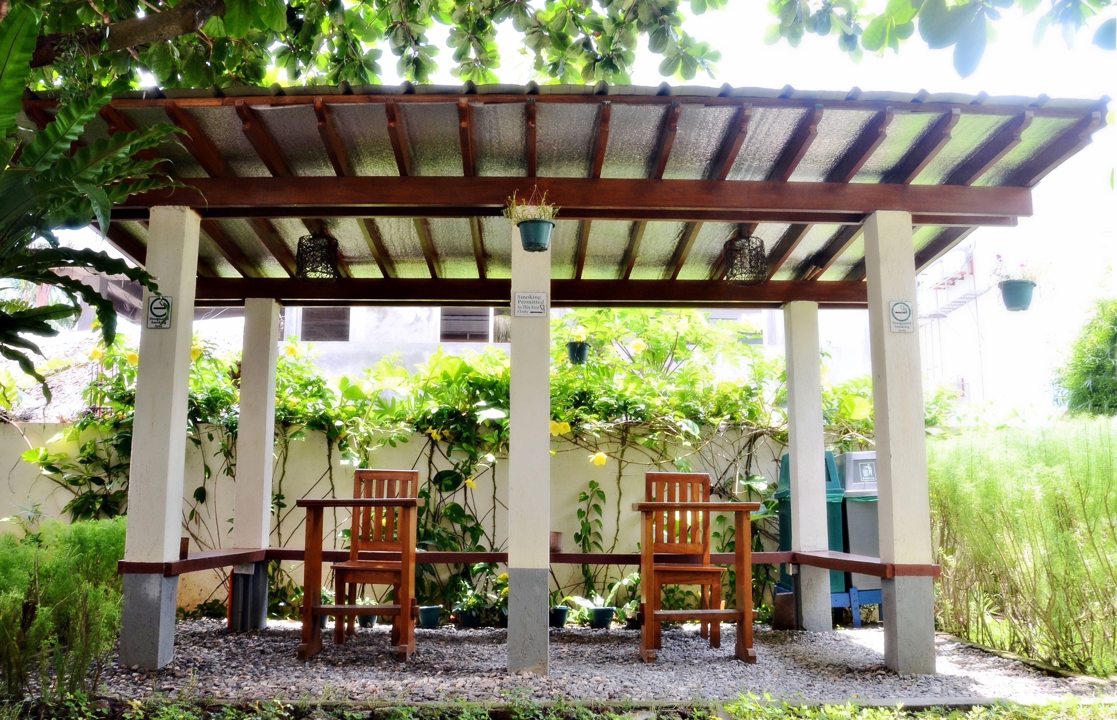 A covered outdoor seating area with two wooden chairs and a table, surrounded by greenery and a pergola structure.