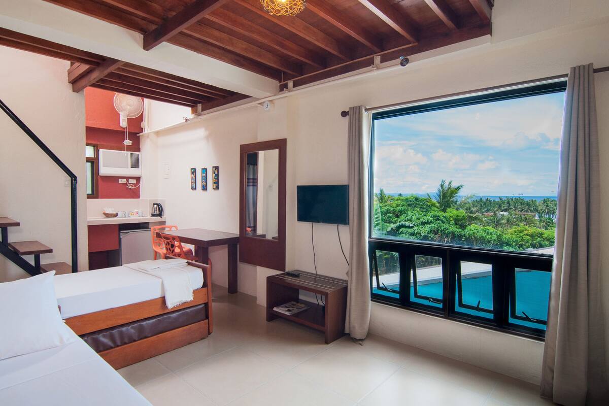 Habitación de hotel con cama, escritorio, televisor y ventana grande con vistas a zonas verdes y piscina.