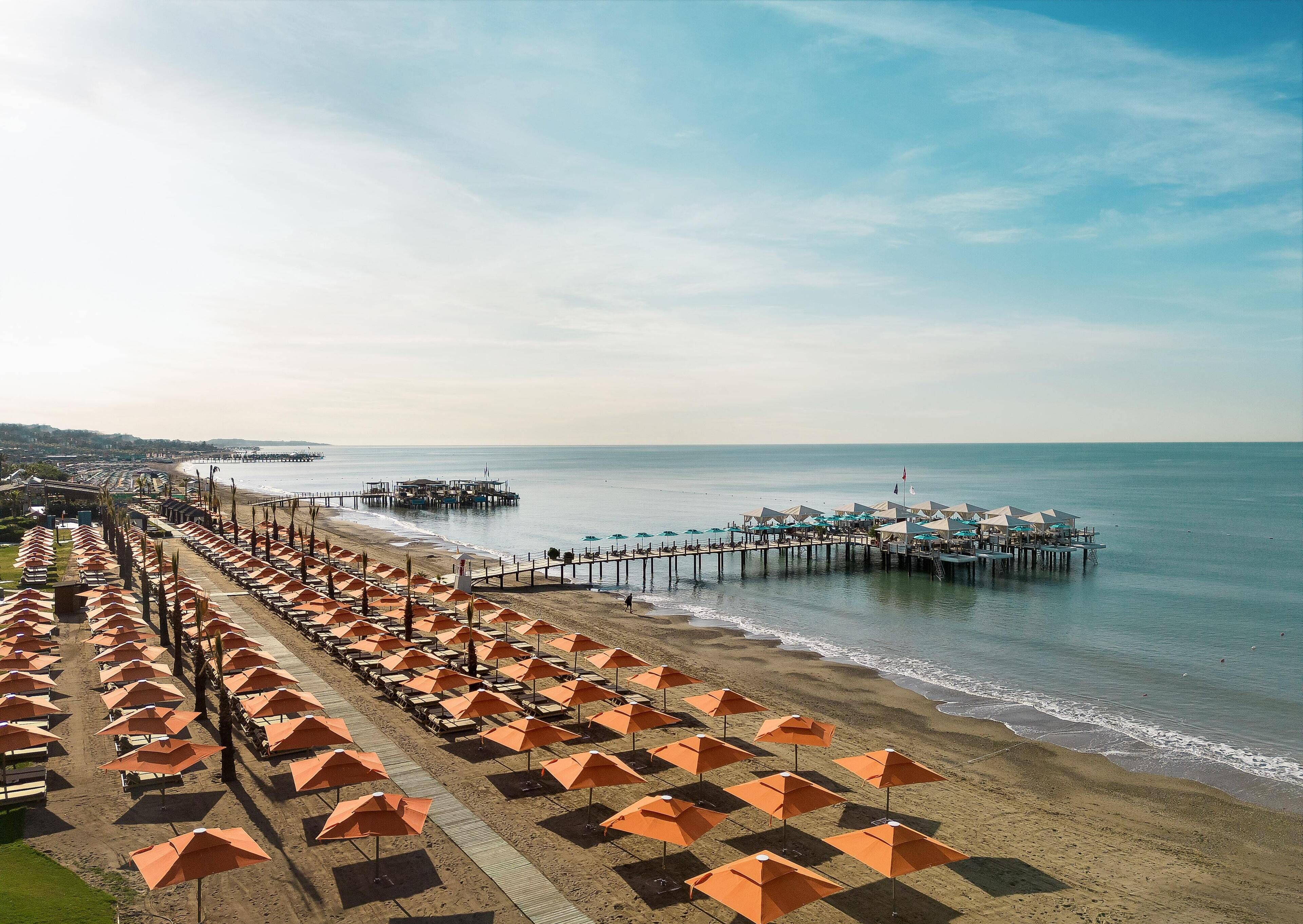 Ein Strand mit Reihen orangener Sonnenschirme und Liegestühle, eine Pier, die ins Wasser ragt, und ein weiter Blick auf eine Stadtkulisse.