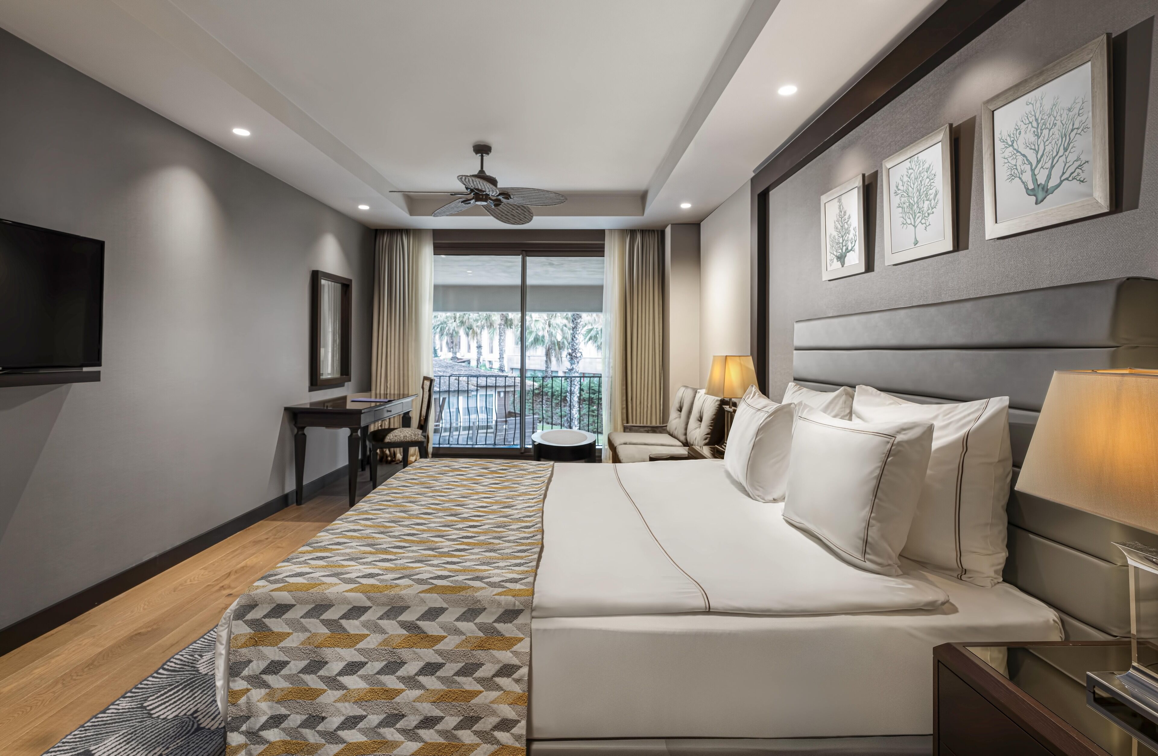 A modern hotel room with a large bed, a desk, a chair, and a balcony with a view of palm trees.