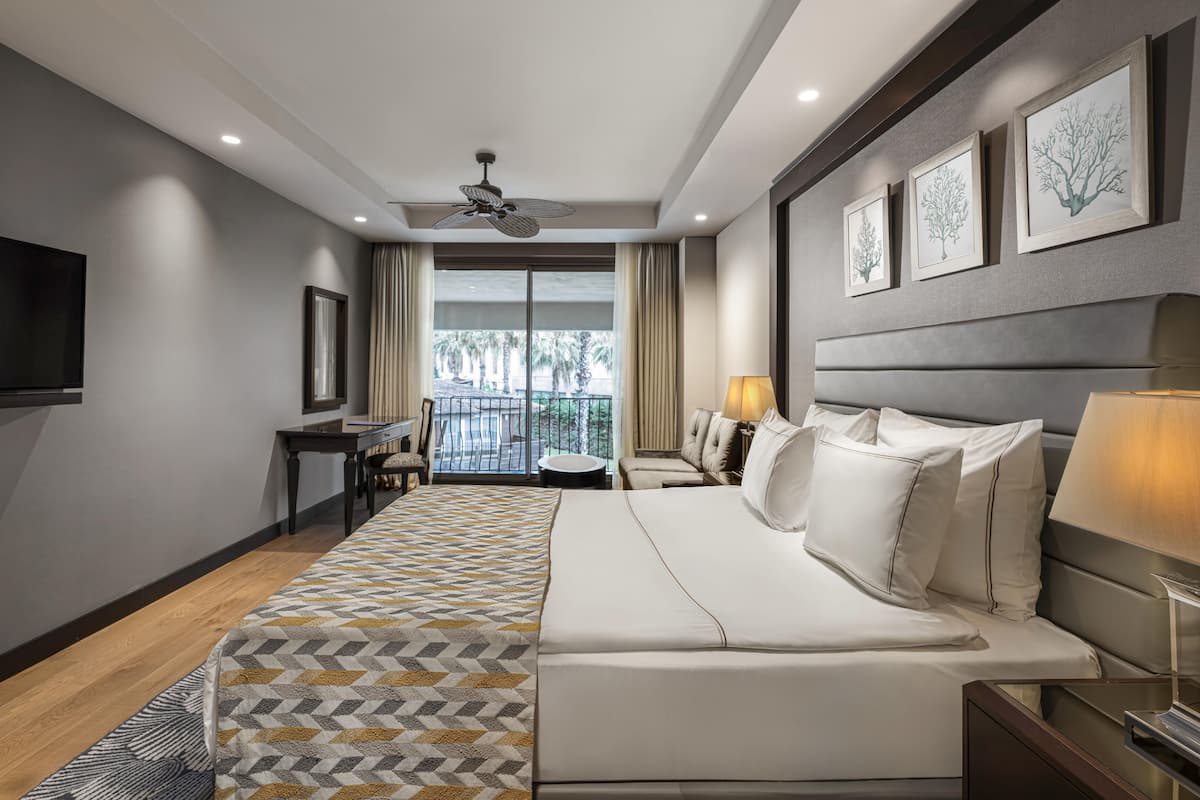 A modern hotel room with a large bed, a desk, a chair, and a balcony with a view of palm trees.
