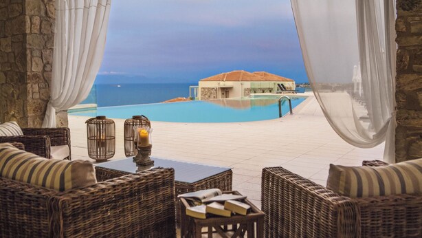 Un espace aménagé au bord de la piscine, avec des meubles en osier, une table en verre et une vue sur la mer et un bâtiment en arrière-plan.
