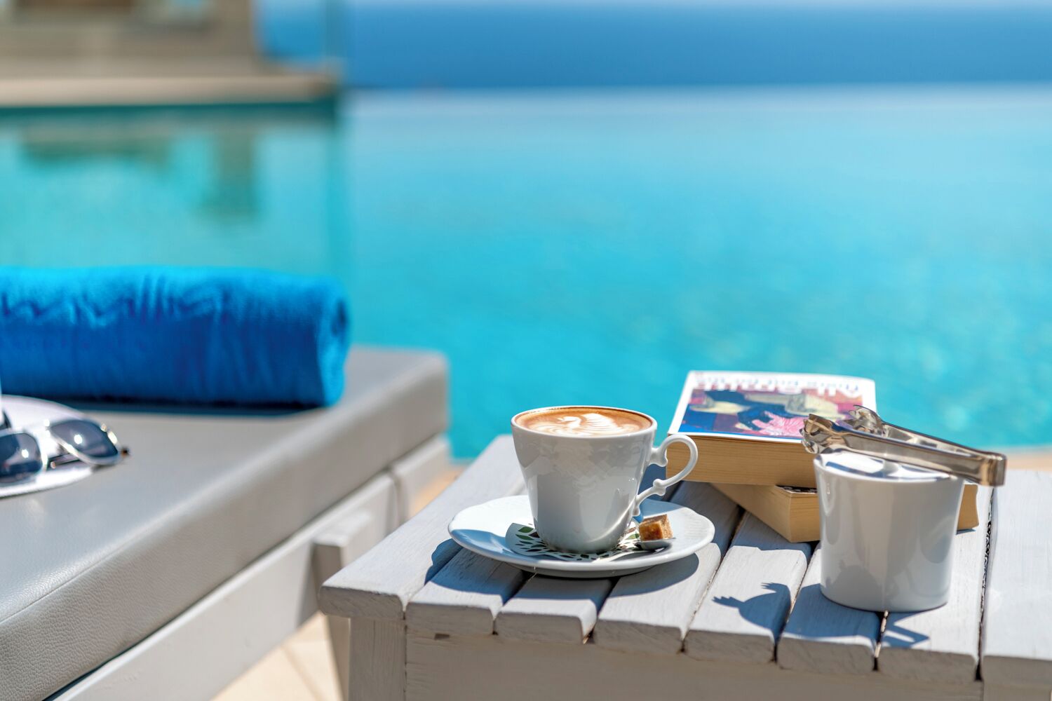 A poolside setting with a cup of coffee, sunglasses, and a book on a wooden table.
