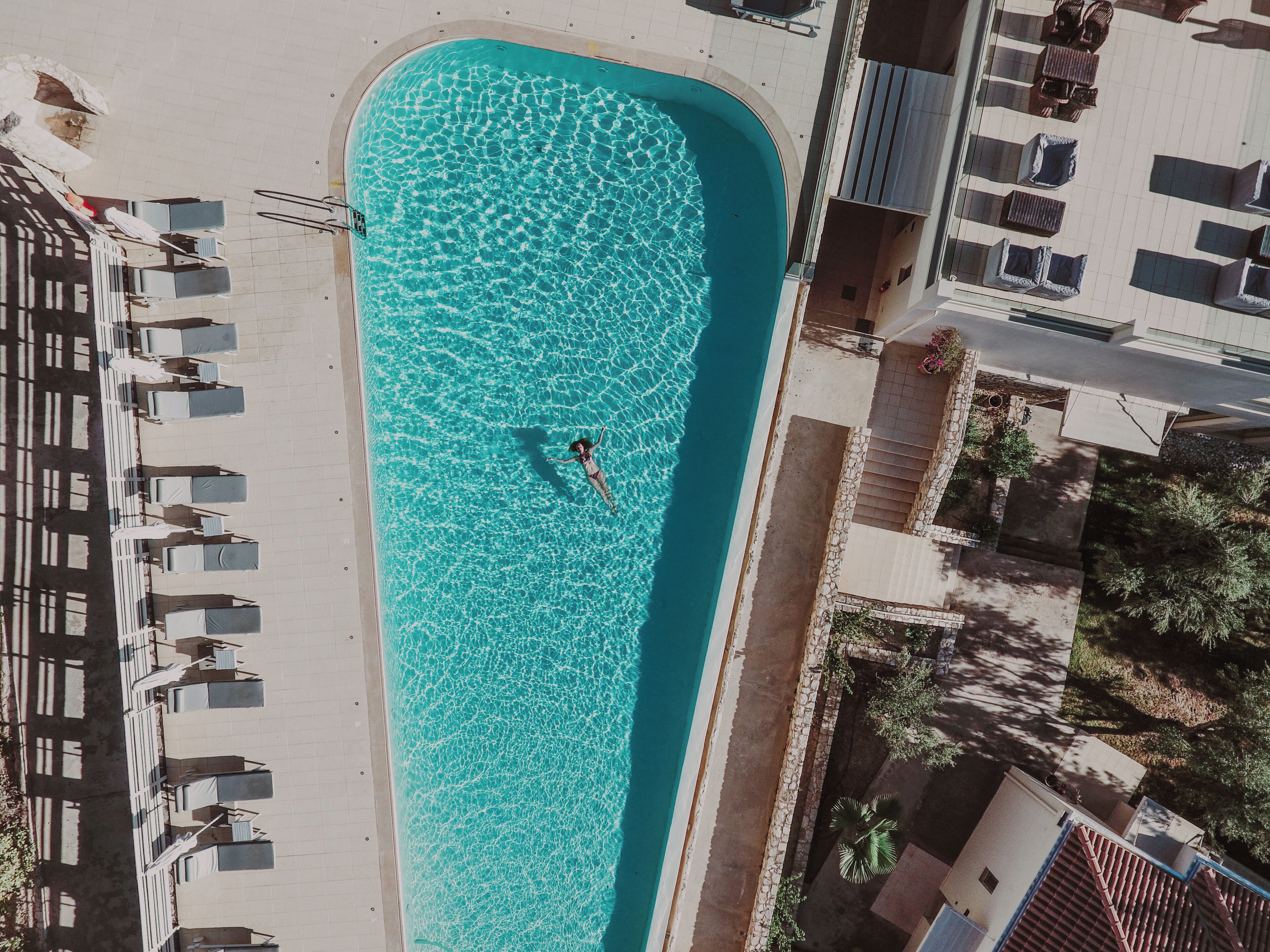 An aerial view of a swimming pool with a person in the water, surrounded by lounge chairs and a building with a tiled roof.