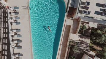 An aerial view of a swimming pool with a person in the water, surrounded by lounge chairs and a building with a tiled roof.