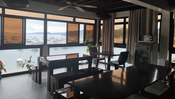 Un espace repas moderne avec une grande table et des chaises, de grandes fenêtres offrant une vue sur la mer et les montagnes, et un ventilateur de plafond.