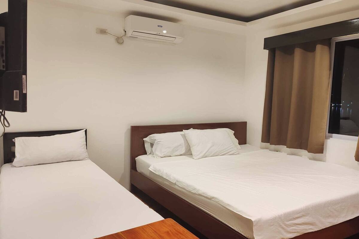 A hotel room with a bed, a bedside table, an air conditioning unit, and a window with curtains.